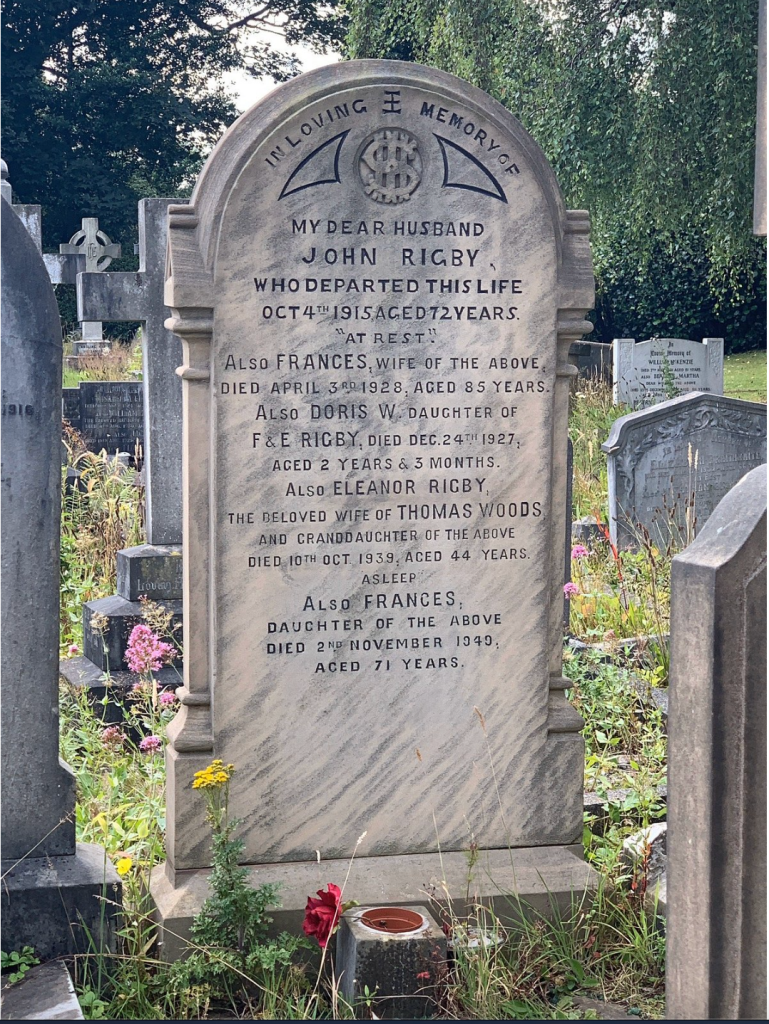 A grave marker inscribed with names and dates, including 'John Rigby' and 'Eleanor Rigby,' surrounded by greenery and flowers.