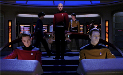 A scene from a science fiction series featuring three characters in Starfleet uniforms on a spaceship bridge. Two characters are seated at control panels, one in a red uniform and the other in a gold uniform, while a standing character in a red uniform serves as the commanding officer. The bridge is illuminated by blue lighting with screens and buttons in the background.